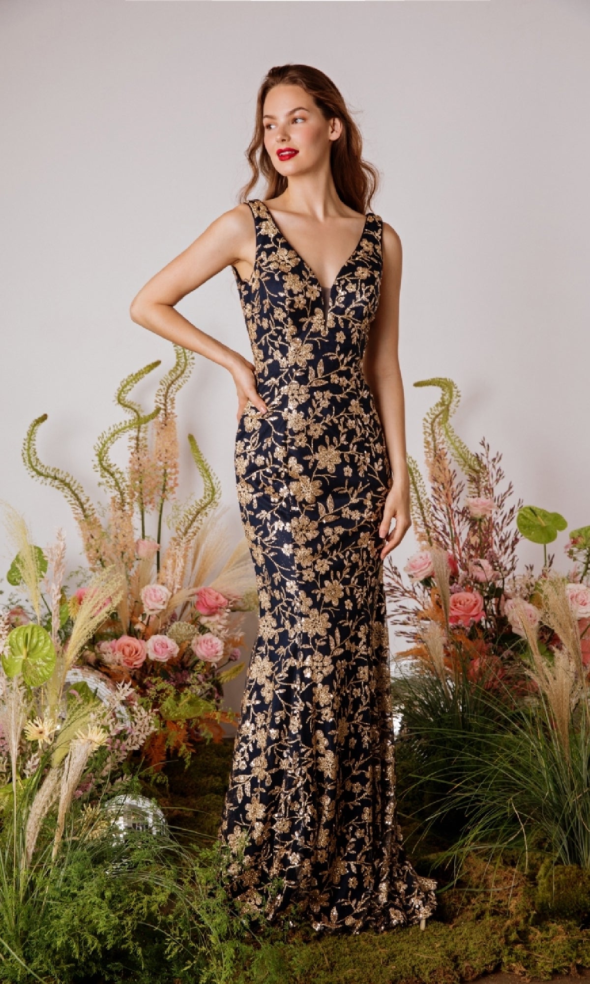 A woman stands before a floral display, wearing the Eureka 9706 Sequin-Print Long Prom Dress—a sleeveless, floor-length formal gown featuring gold sequin floral accents.